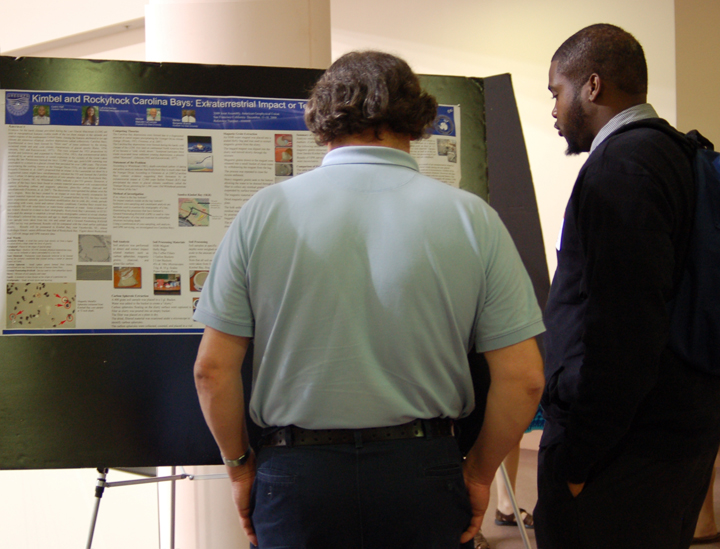 Two men stand in front of a research project poster